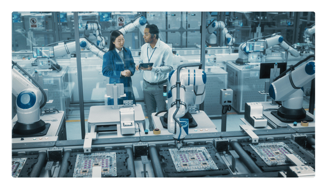 Two manufacturing leaders review production data on a tablet beside robotic arms on an automated circuit board assembly line.