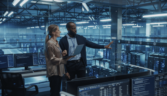 Two team members review information on a laptop in a control room overlooking rows of server racks.