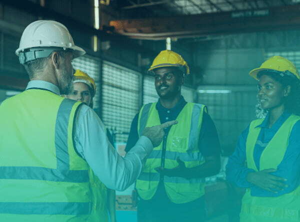 A manufacturing supervisor in a hard hat and safety vest talks with three team members on the plant floor.