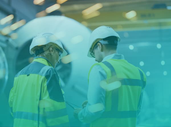 Two manufacturing professionals in hard hats and safety vests review a tablet together on a plant floor beside large industrial equipment.